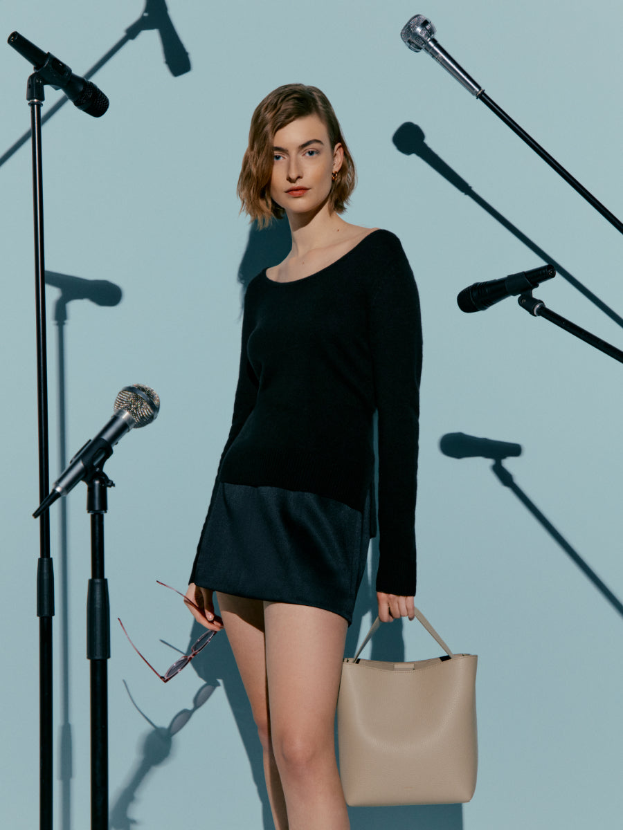 Woman standing among multiple microphones holding a bag and glasses.