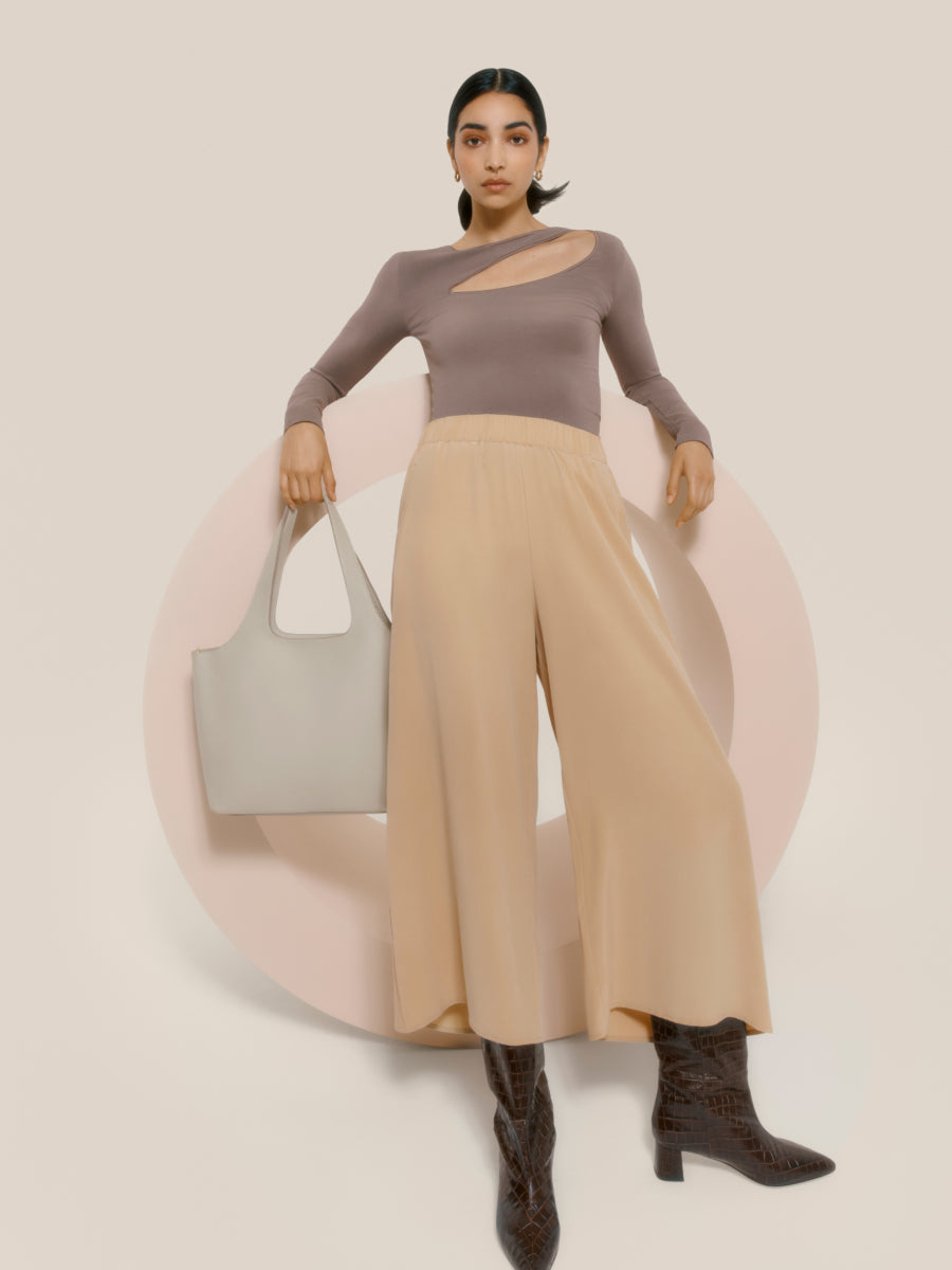 Woman standing with a handbag and wearing a long-sleeve top and wide-leg pants.