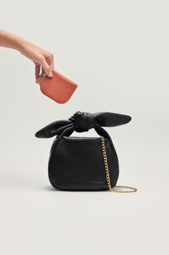 Hand placing a wallet into a handbag with a knotted handle and chain strap on a plain background.