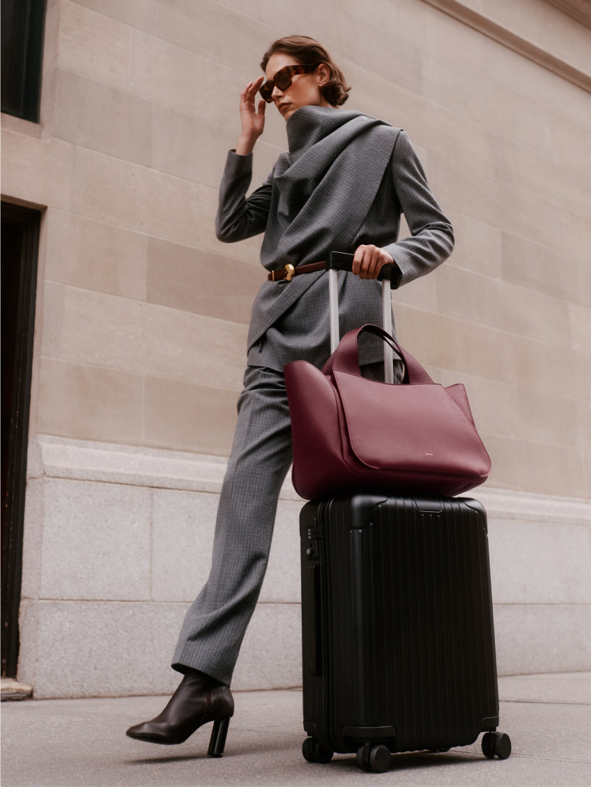 Person in a suit with a travel bag and suitcase, hand adjusting sunglasses.