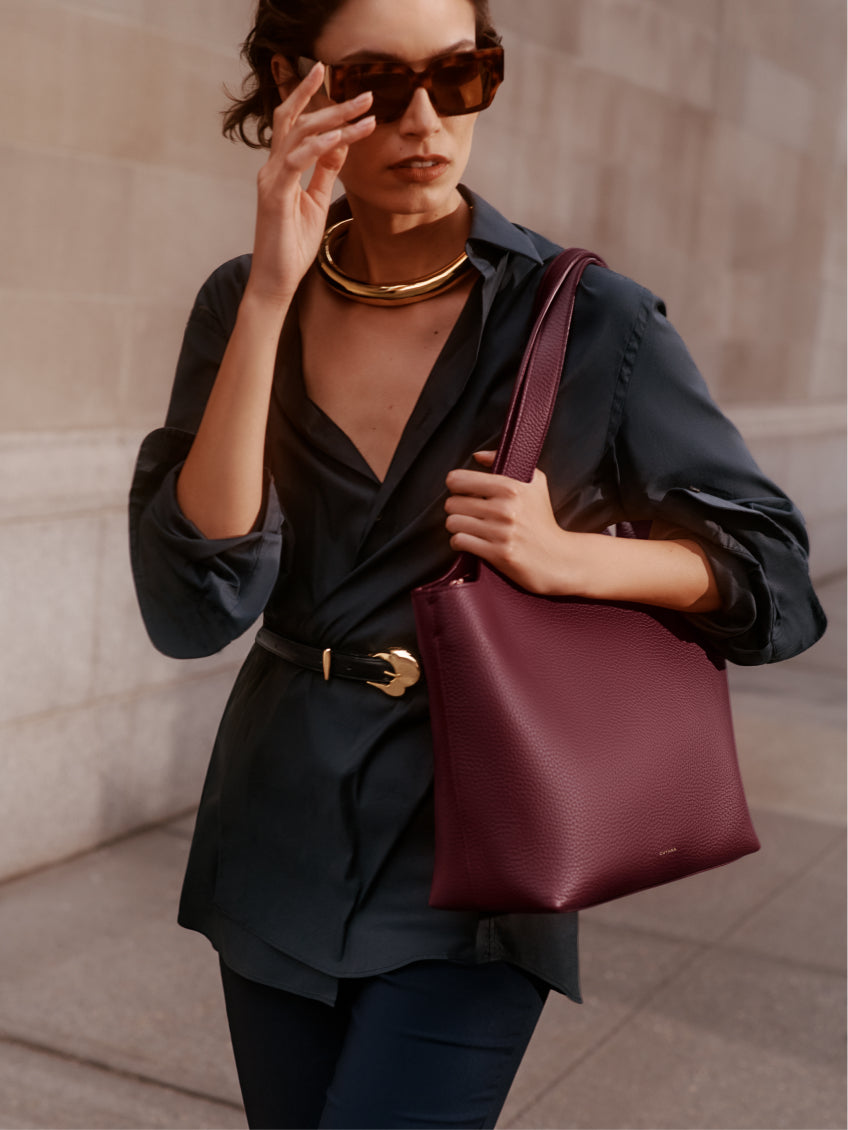 Person wearing sunglasses, holding a shoulder bag and adjusting glasses with left hand, wearing a belt.