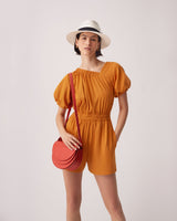 Woman in a romper and hat standing with one hand on hip and holding a round bag.