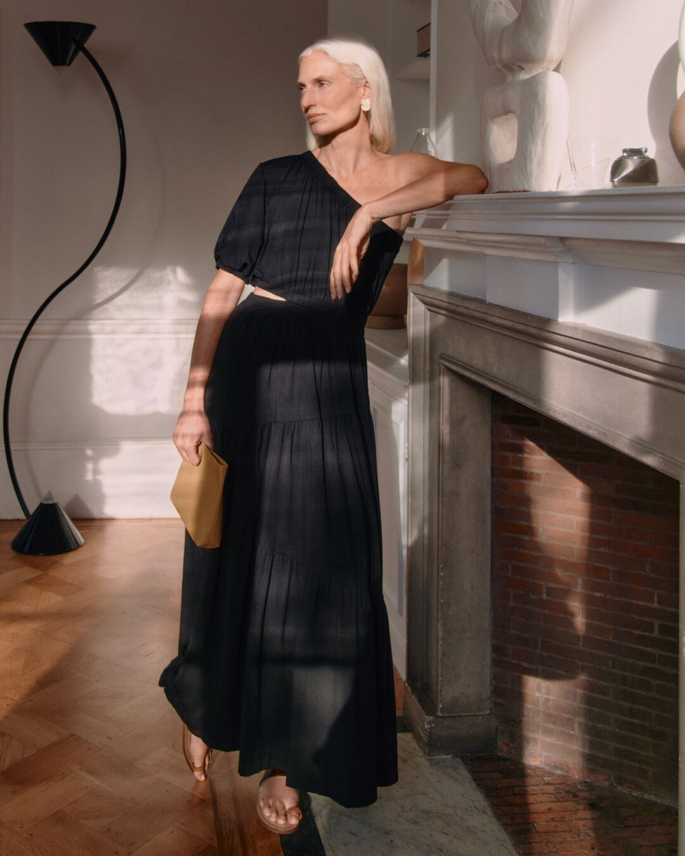 Woman standing by fireplace in elegant dress, holding a clutch.
