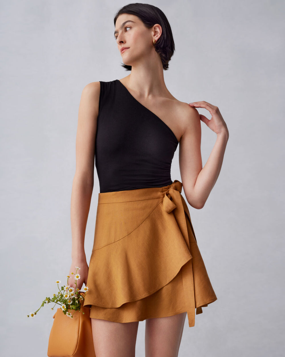 Woman standing in asymmetrical top and wrap skirt, holding a small bag with flowers.