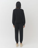 Woman standing, facing away, wearing a long-sleeve top and pants with sneakers.