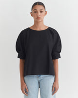 Woman in a blouse and jeans standing against a plain background.