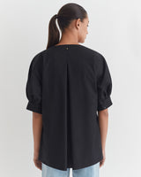 Woman standing, viewed from the back, wearing a buttoned shirt and jeans.