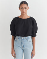 Woman in a blouse and jeans standing with hands at sides.