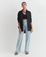 Woman standing in an open shirt, crop top, jeans, and sandals.