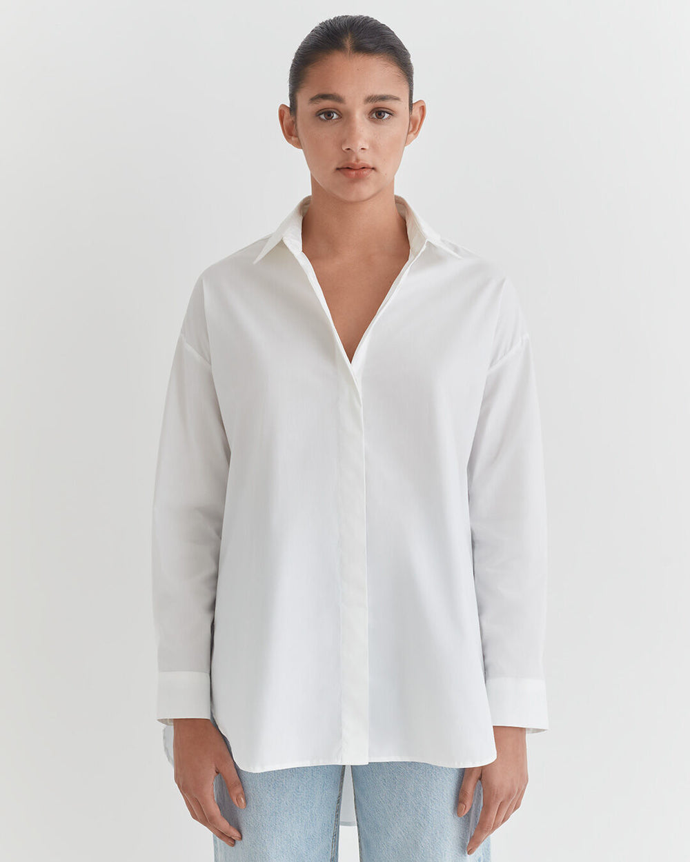 Poplin Oversized Shirt Woman in a button-down shirt and jeans standing against a plain background.