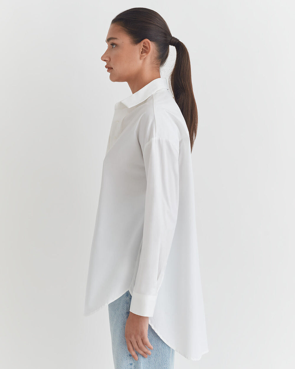 Poplin Oversized Shirt Woman standing in profile with a ponytail, wearing a shirt and jeans.