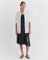 Woman standing in a dress with a button-up shirt over it and sandals.