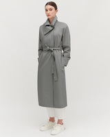 Woman standing in a coat with belt, wearing trousers and sneakers.