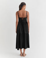 Woman standing facing away, wearing a mid-length dress with straps.