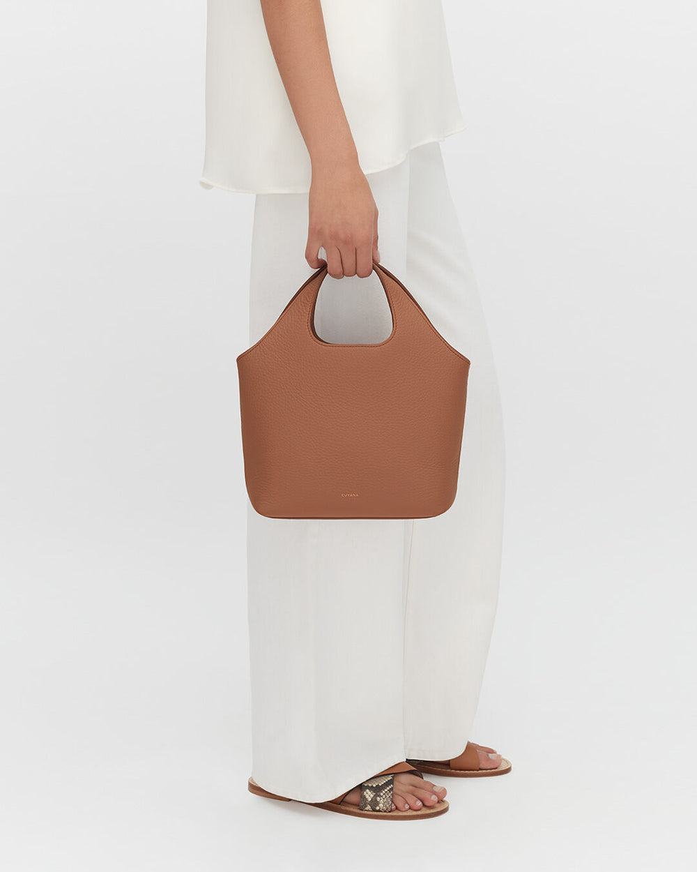 Person standing and holding a handbag while wearing pants and sandals.