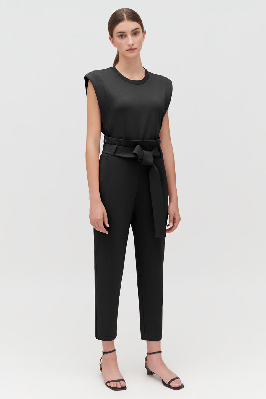 Woman standing in a sleeveless top and belted pants with sandals.