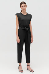 Woman standing in a sleeveless top and belted pants with sandals.