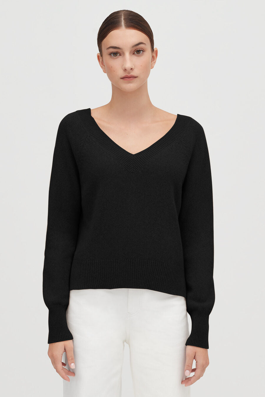 Woman standing with hands slightly apart, wearing a V-neck sweater and pants.