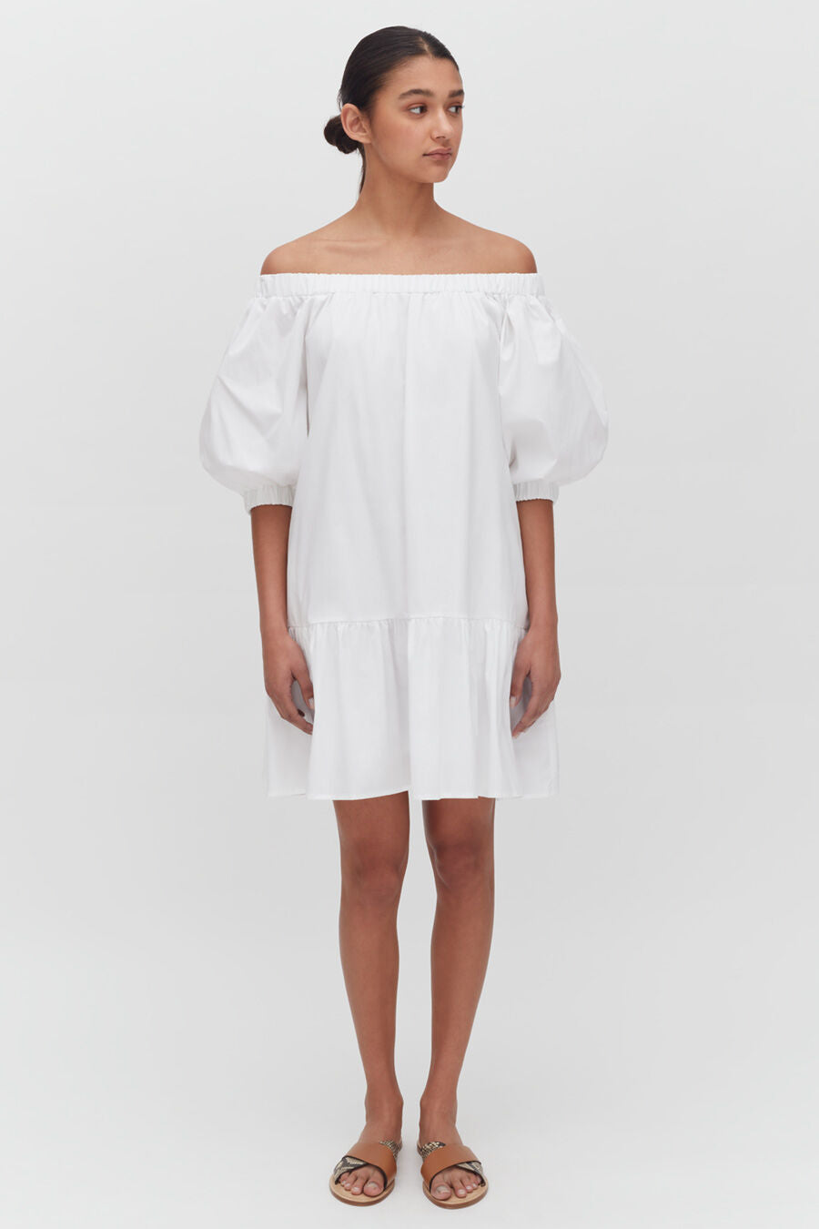 Woman in off-shoulder dress and sandals standing against a white background.