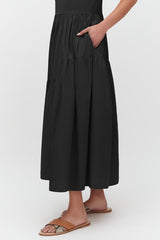 Person standing in a long skirt with hand in pocket, wearing sandals.