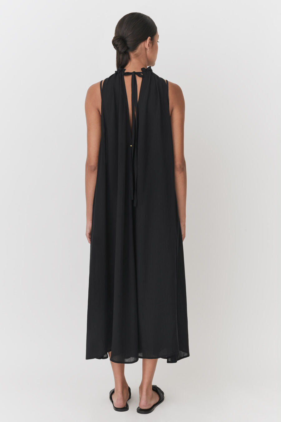 Woman standing facing away, wearing a long sleeveless dress and sandals.