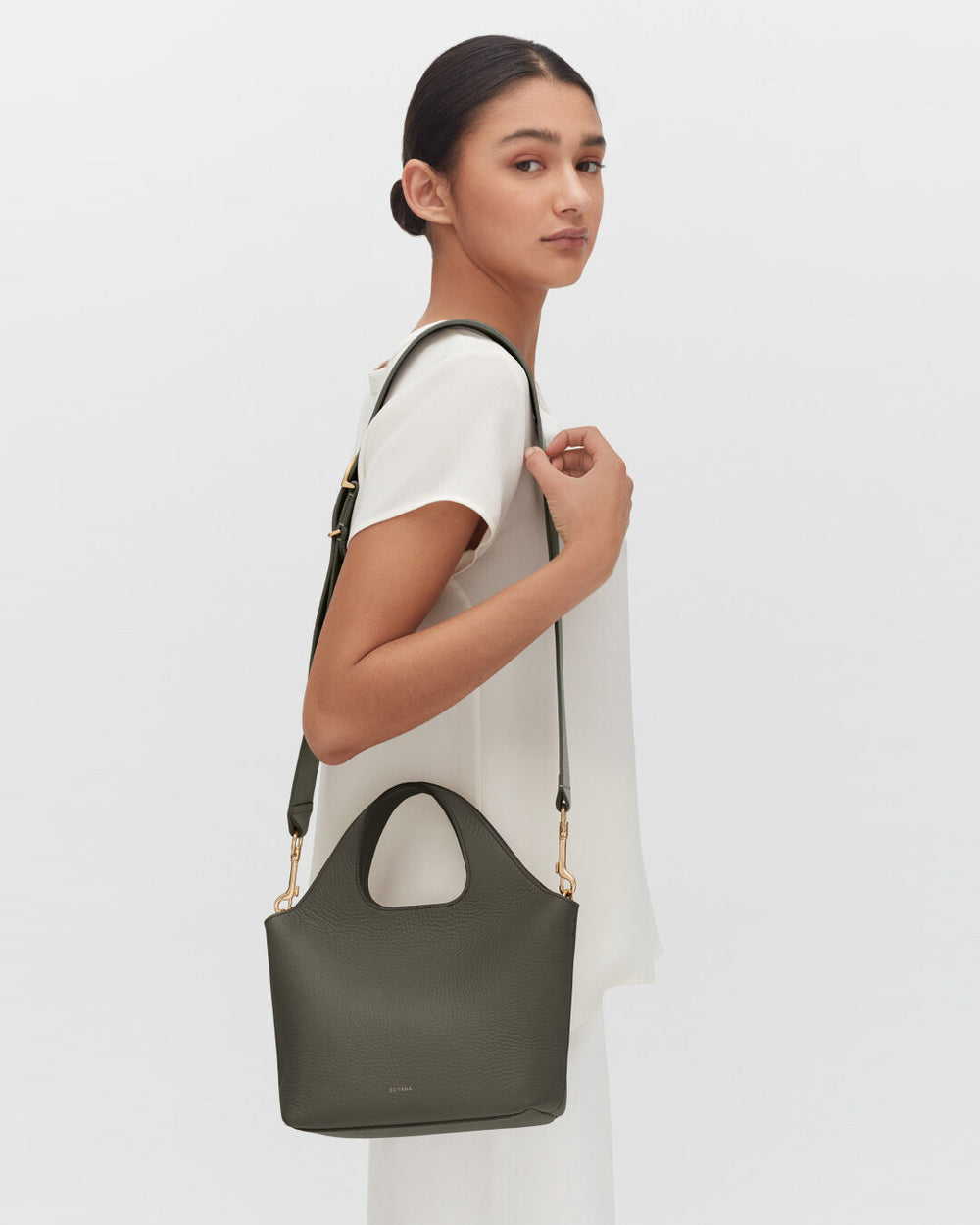 Woman standing sideways holding a handbag on her shoulder.