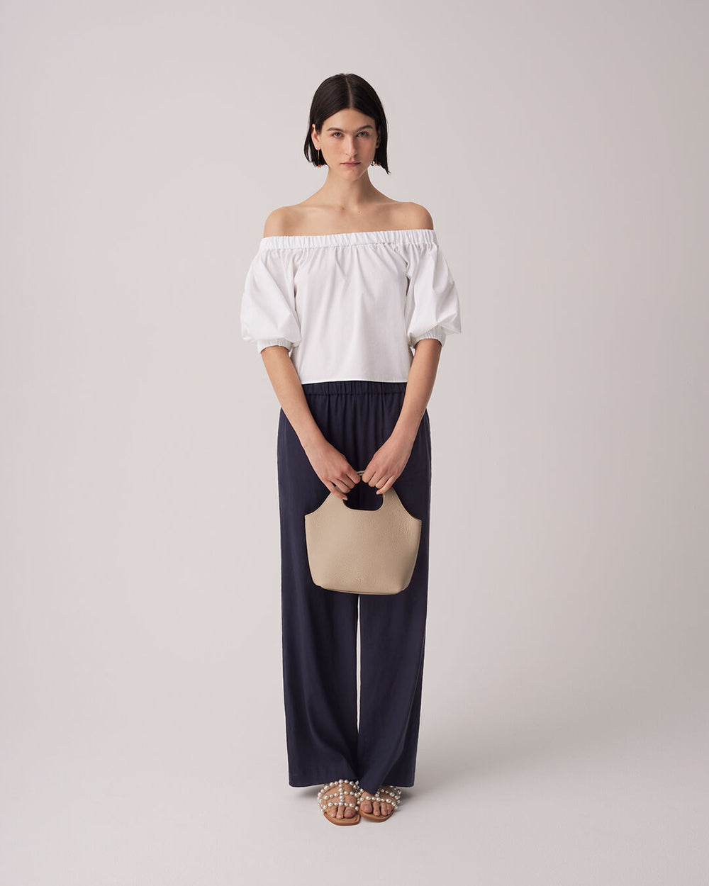 Woman in off-shoulder top and pants holding a purse, standing upright.
