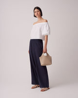Woman standing in off-shoulder top, wide pants, holding a bag.