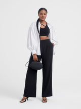 Woman standing in a studio with jacket, crop top, pants, heels, and a handbag.