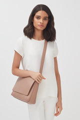 Woman standing, wearing a top and trousers, holding a shoulder bag.