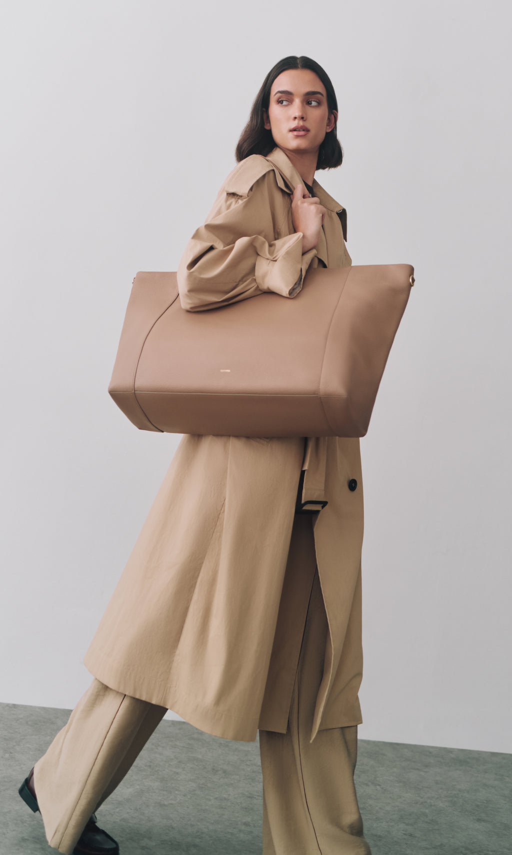 Model walking with large travel bag and wearing a long coat.