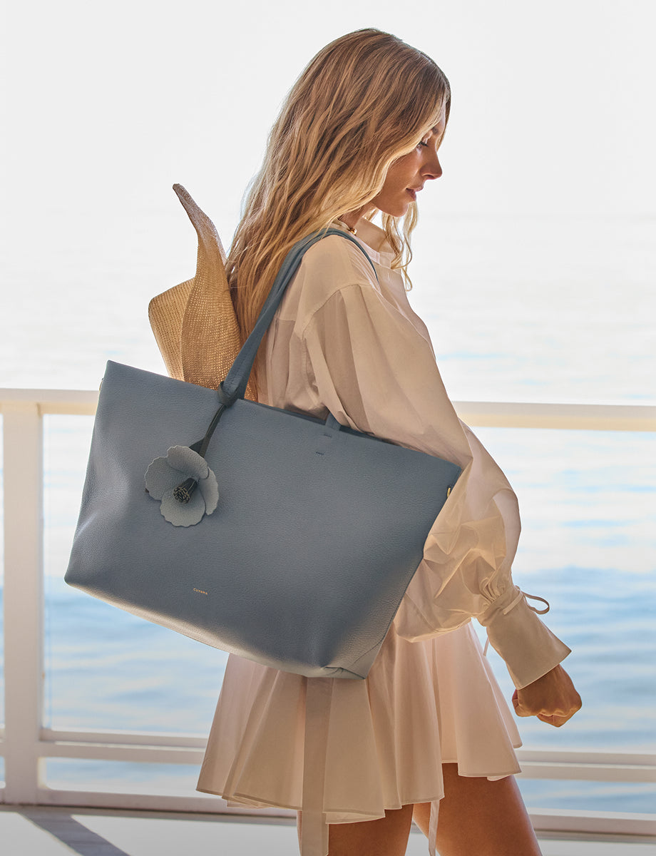 Person holding a tote bag with a flower ornament on a balcony facing the ocean.