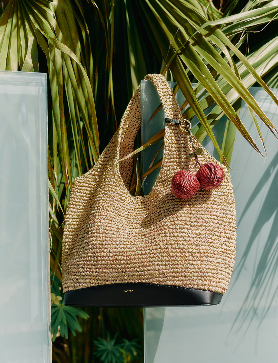 Woven handbag with pom-poms, hanging in front of palm leaves.