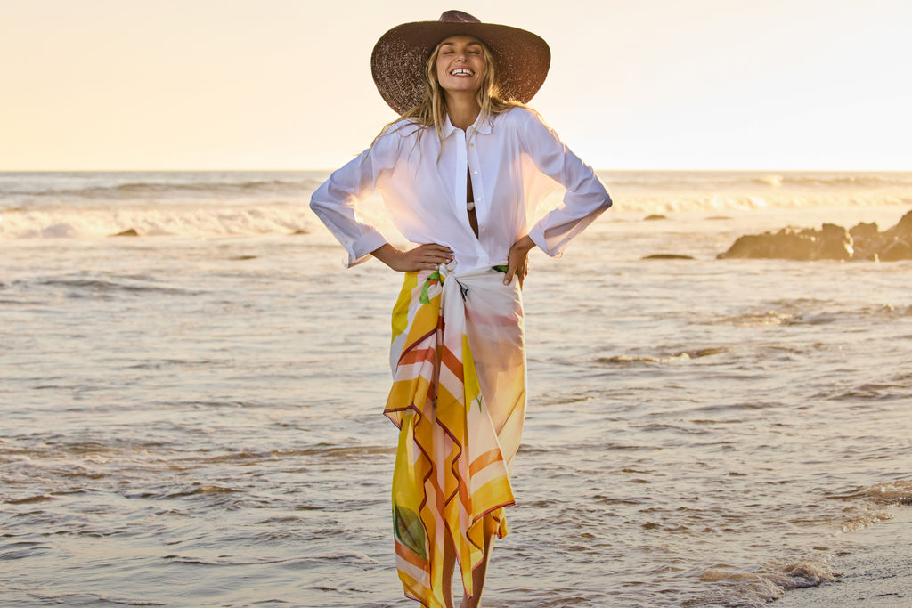 Person in wide-brimmed hat and shirt standing on a beach with the ocean in the background.