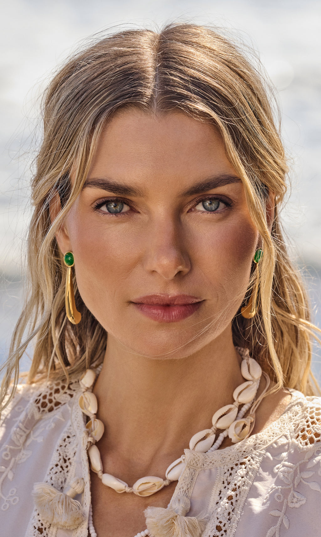 Person wearing earrings and a shell necklace by water.