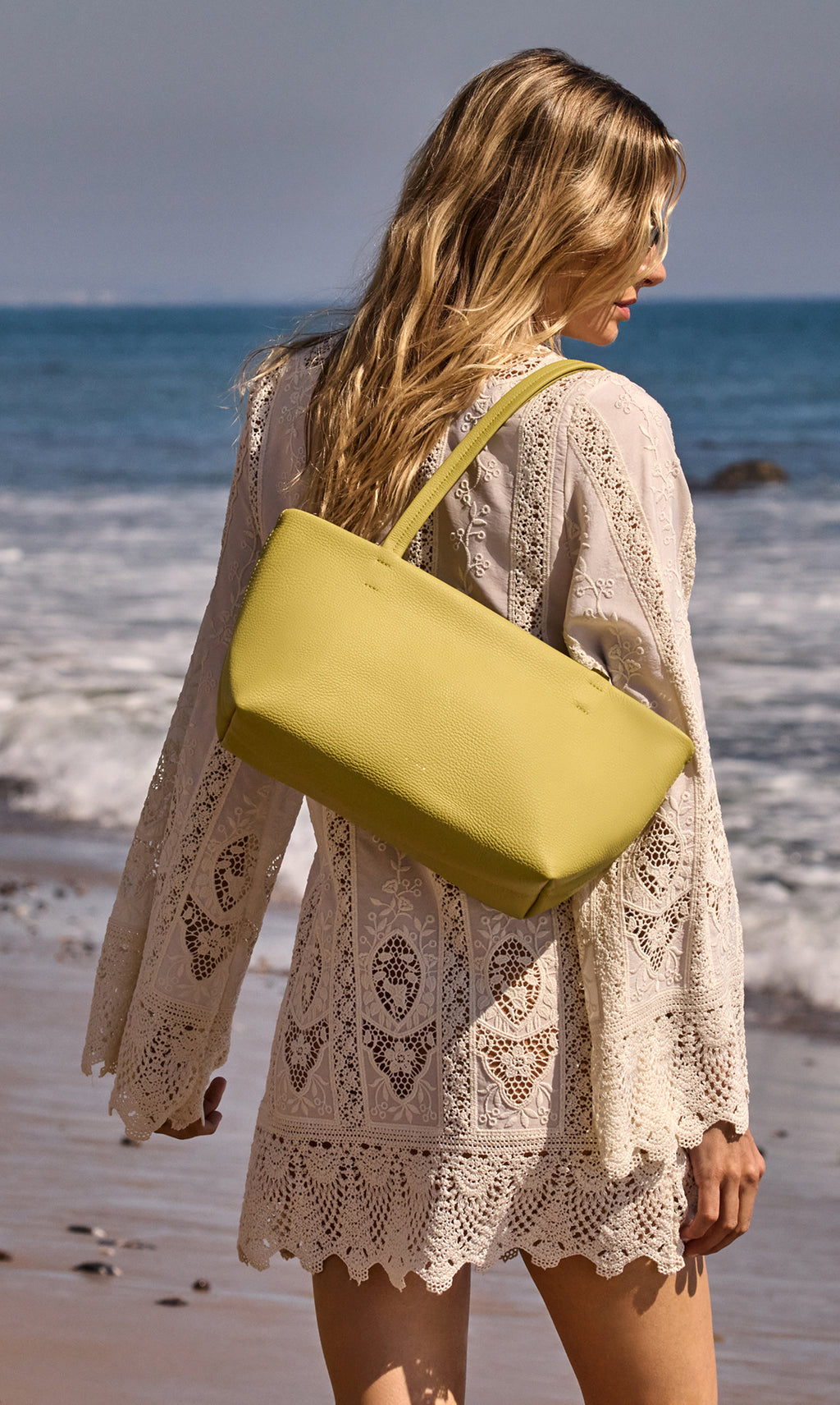 Woman by the ocean wearing a lace dress and carrying a handbag over her shoulder.