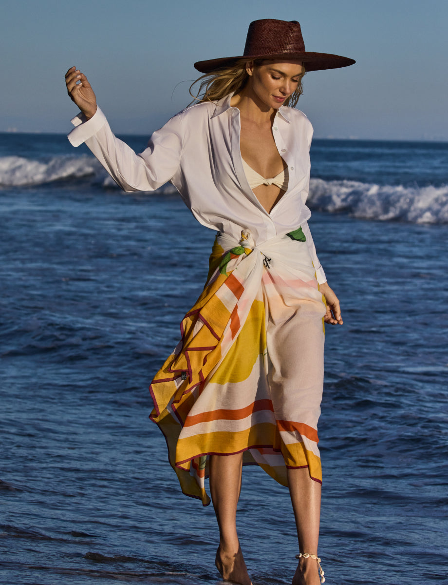 Person in open shirt and skirt stands on the beach near the water.