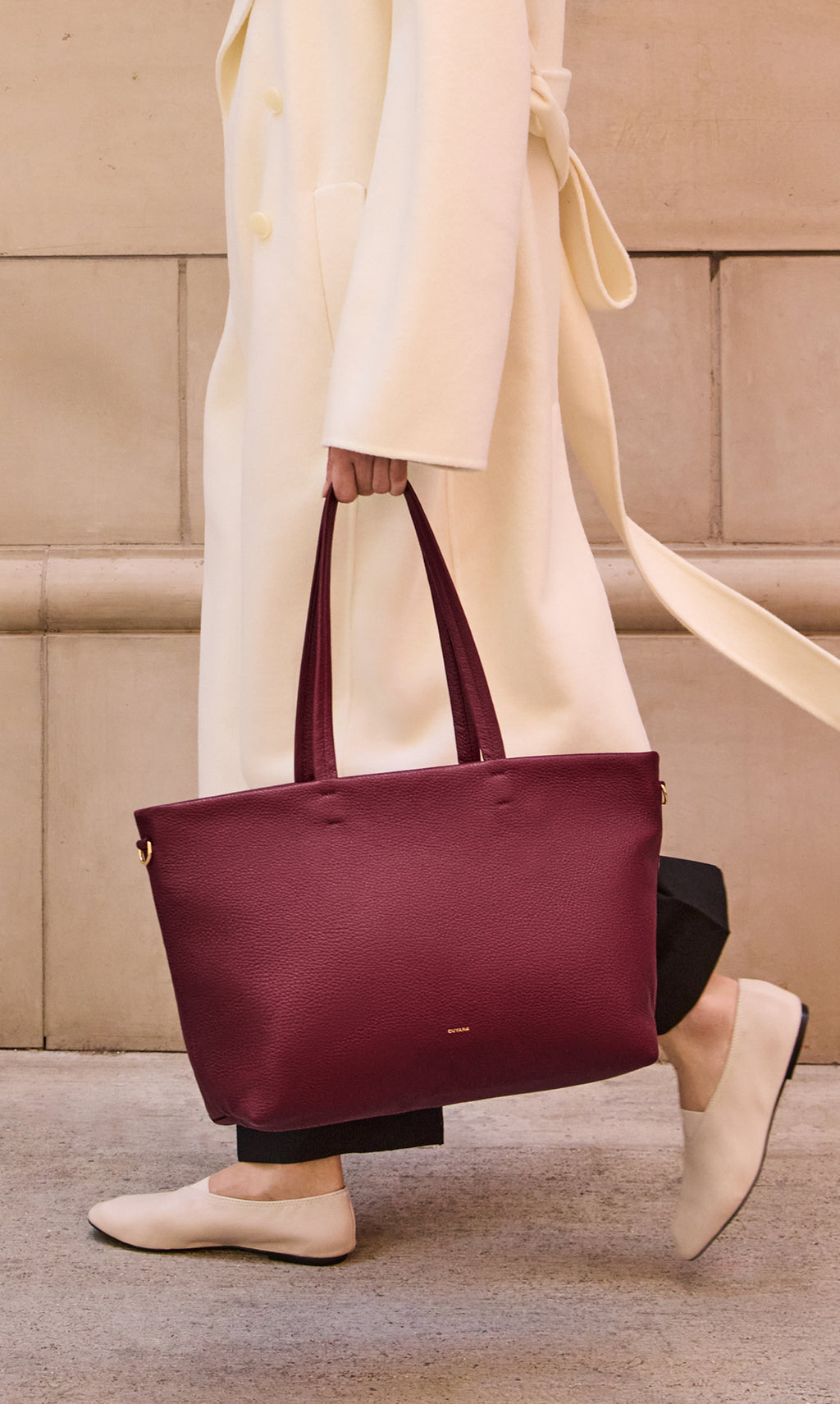 Person with large handbag wearing a long coat walking on a sidewalk.