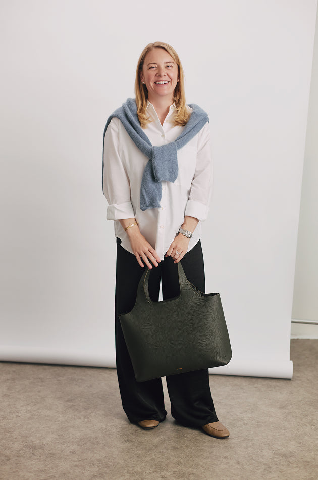 Person smiling, wearing a sweater over shoulders, holding a large tote bag in hand.