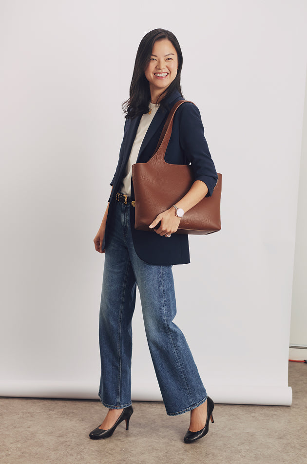 Person smiling, wearing a blazer, jeans, heels, and holding a large shoulder bag