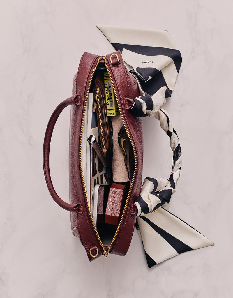 Open handbag with scarf handle showing items like a book and lipstick inside.