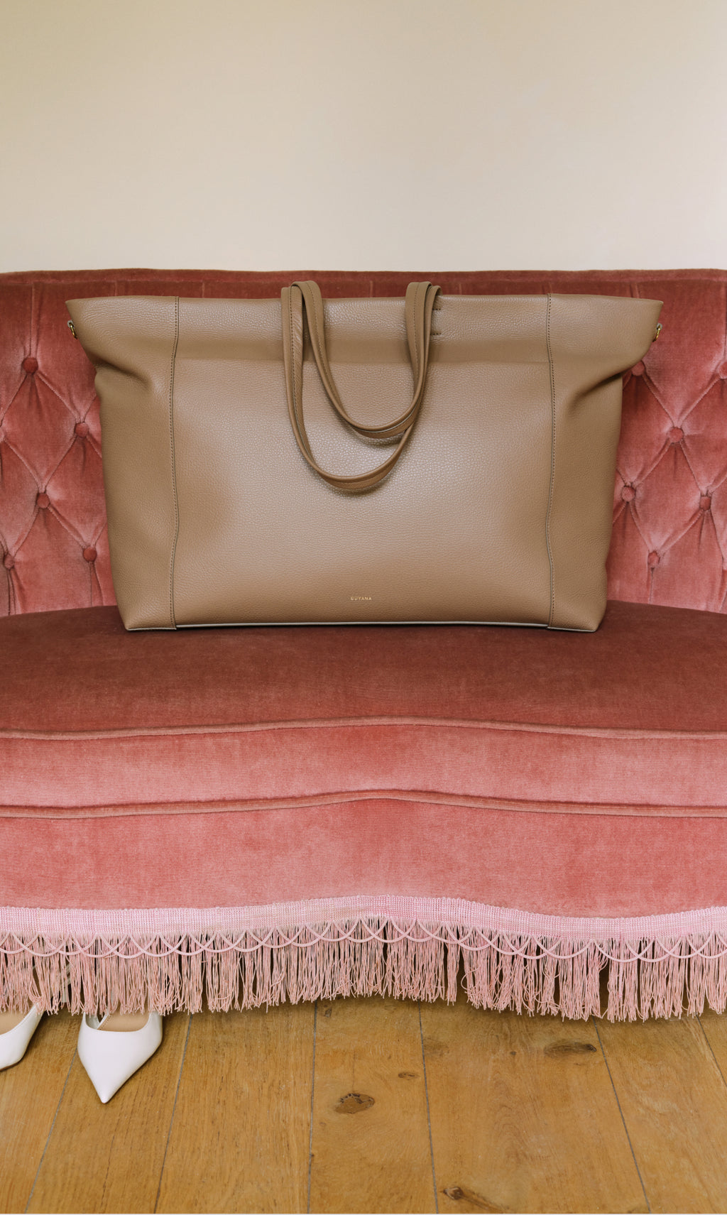 Large travel bag on a tufted sofa with fringed trim and shoes on the floor.