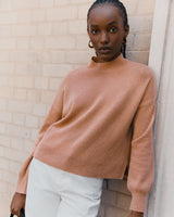 Woman leaning against a wall wearing a sweater and pants.