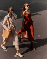 Two women walking on a street, one with sunglasses, both carrying large bags.