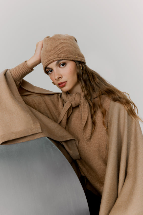 Woman wearing a beanie and cape, leaning on a chair.