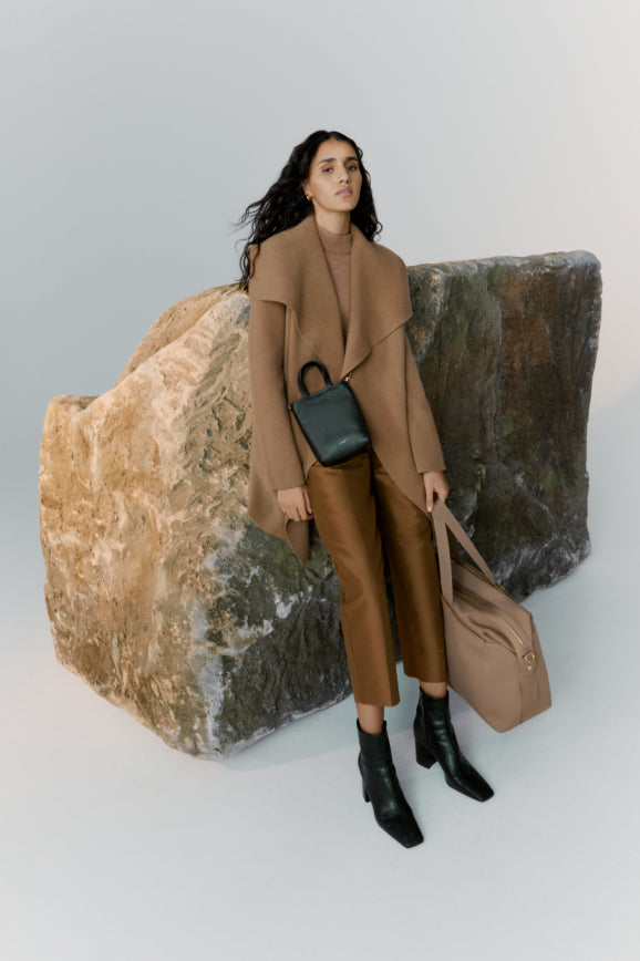 Woman standing next to a large rock, holding a bag with another bag at her side.