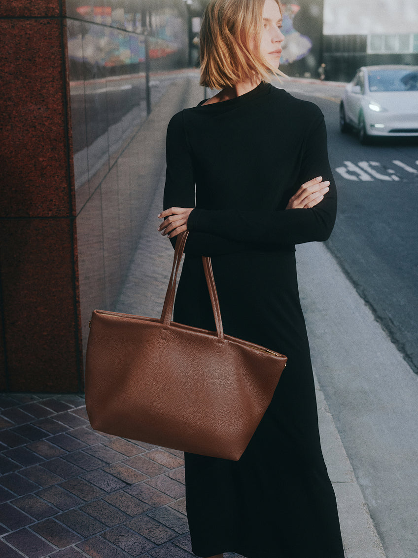 Person in a long dress standing on a sidewalk carrying a large handbag.