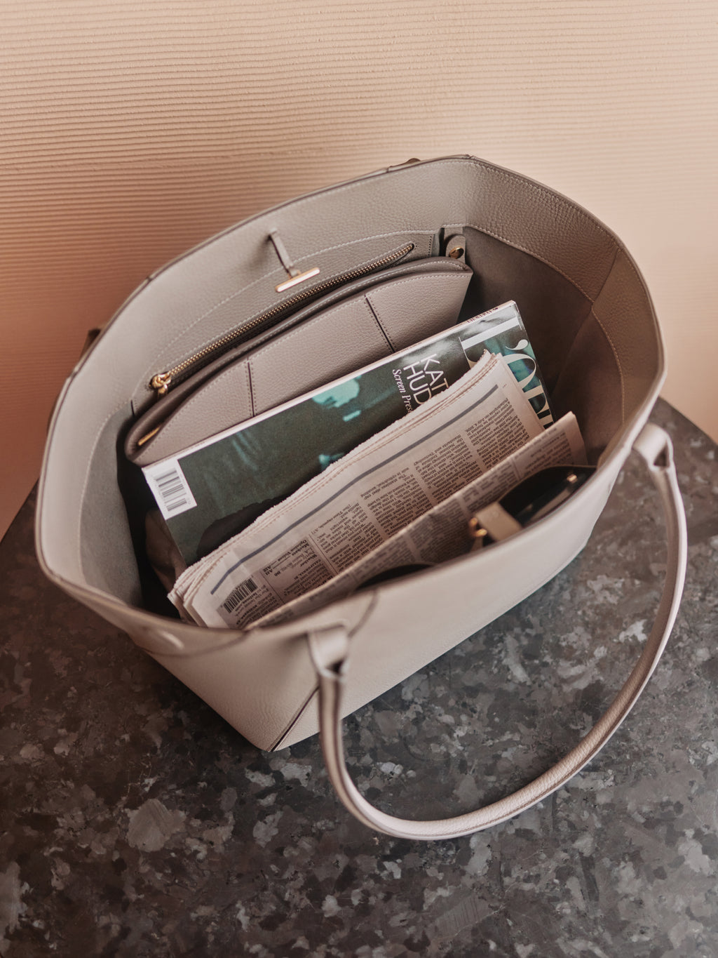 Open tote bag with a newspaper and a book inside on a marble surface.