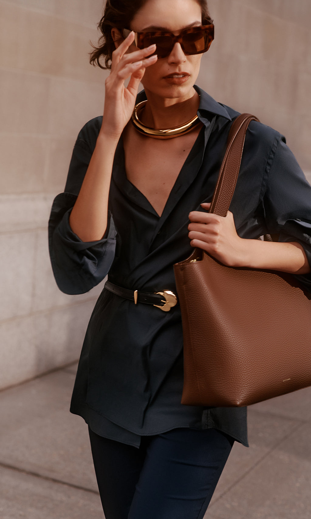 Person in sunglasses holding a shoulder bag, wearing a belt and layered necklace.