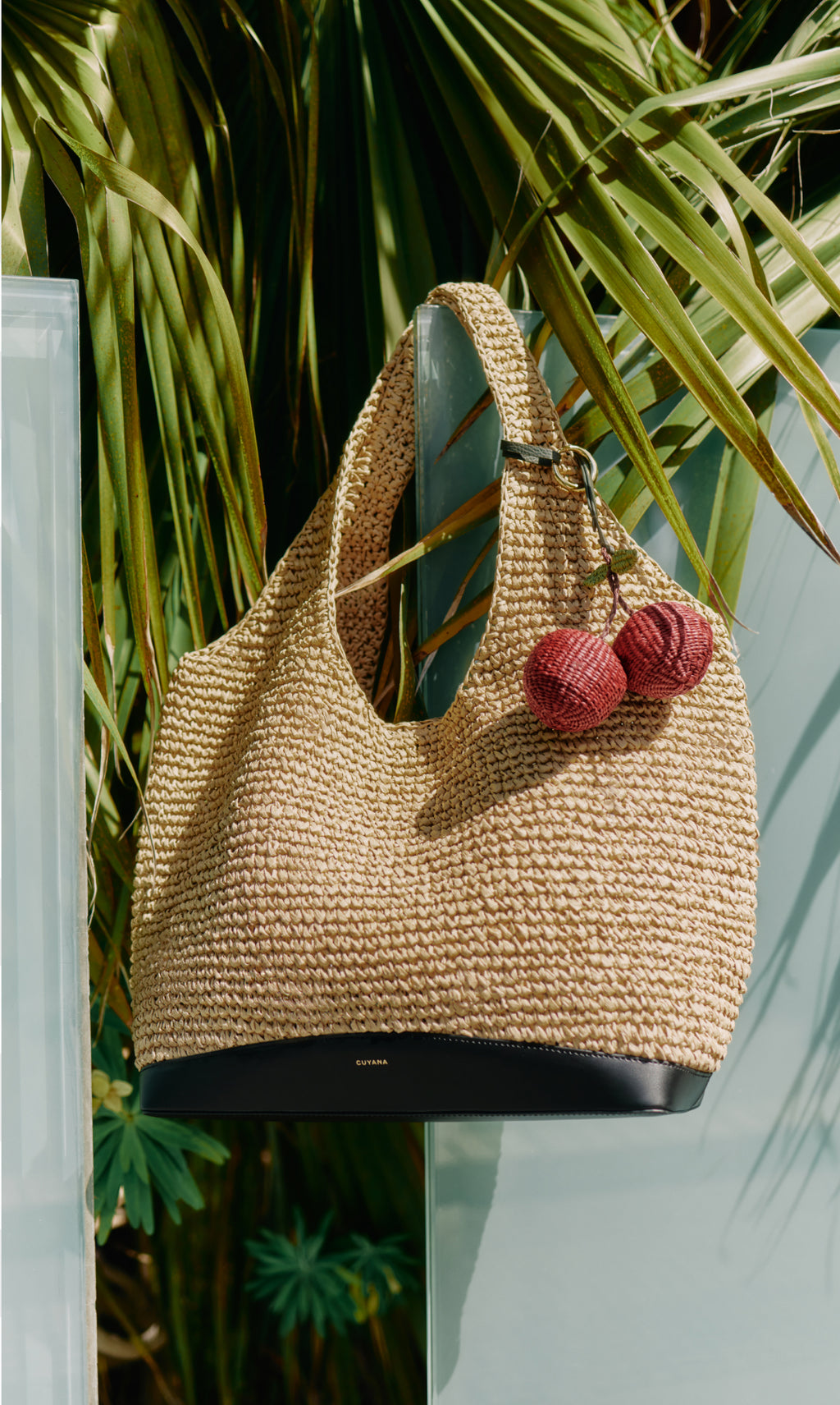 Woven tote bag with pom-pom keychain on glass panel near palm leaves.